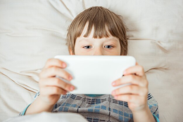 Il piccolo bambino moderno si trova sul letto con lo smartphone nelle sue mani