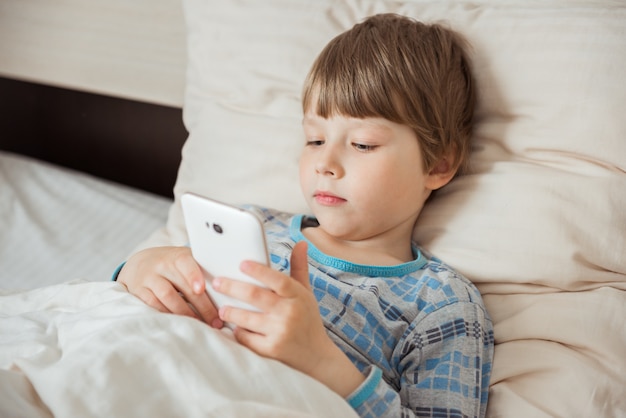 Modern little child lies on bed with smartphone in his hands