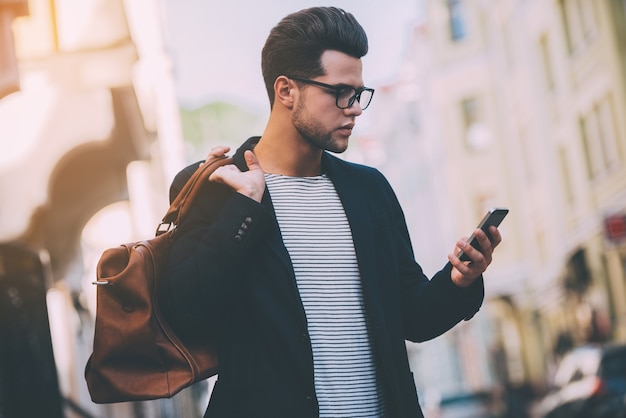 Modern man. Handsome young man in smart casual wear carrying bag on shoulder and looking at his smart phone while standing on the street