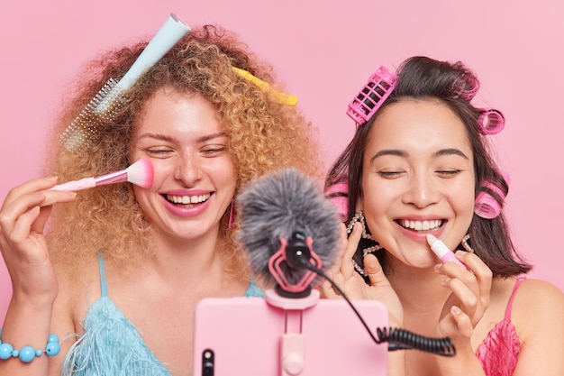 Marketing moderno. donne allegre e diverse registrano contenuti per blog sullo stile di vita applicano cipria e rossetto ridono allegramente danno consigli su come rimanere belle stare uno accanto all'altro davanti alla fotocamera del telefono