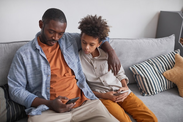 Modern mature man wearing casual clothes sitting on sofa with his teen son showing him photos on smartphone