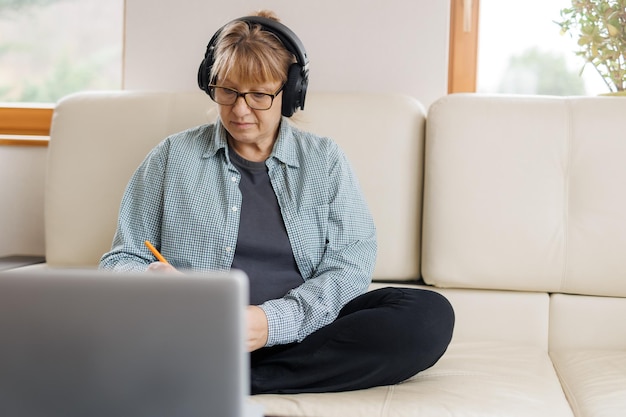 Modern middle aged blond woman in headphones take online course or training on computer at home Smart mature Caucasian female in earphones watch webinar make note study distant on laptop