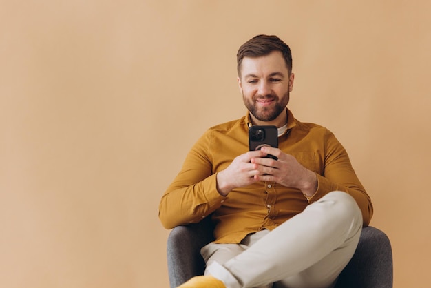 Modern millennial bearded man is happy and smiling in a yellow shirt typing something in smartphone