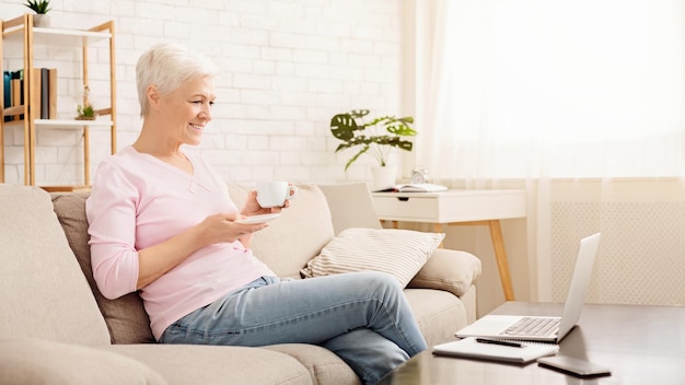 modern morning positive senior woman enjoying coffee and watching news on laptop sitting on couch at
