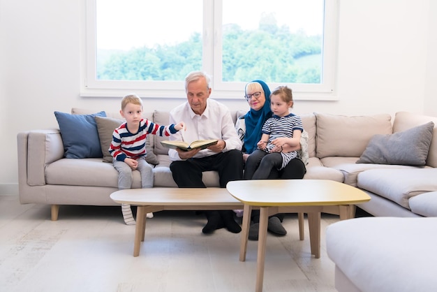 modern muslim family grandparents with grandchildren reading Quran and praying together on the sofa before iftar dinner during a ramadan feast at home