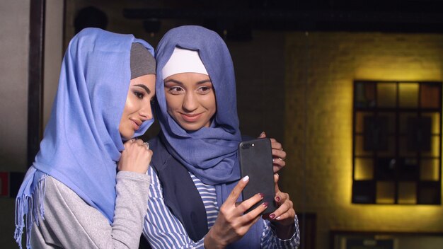 Modern Muslim women take pictures on a mobile phone. Girls in hijabs talking and smiling.