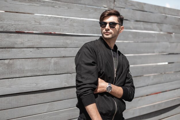 Modern nice young man in a fashionable black jacket in stylish sunglasses is posing outdoors near a wooden vintage wall. Attractive guy resting on the street on a sunny summer day.