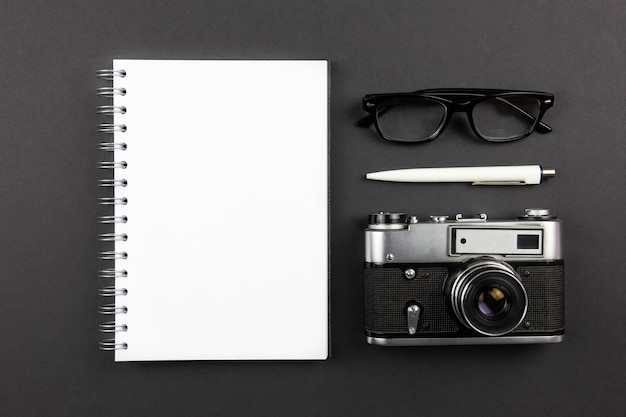 Modern office workplace flat lay with blank spiral paper notebook mockup, retro camera, pen and reading glasses on dark black surface