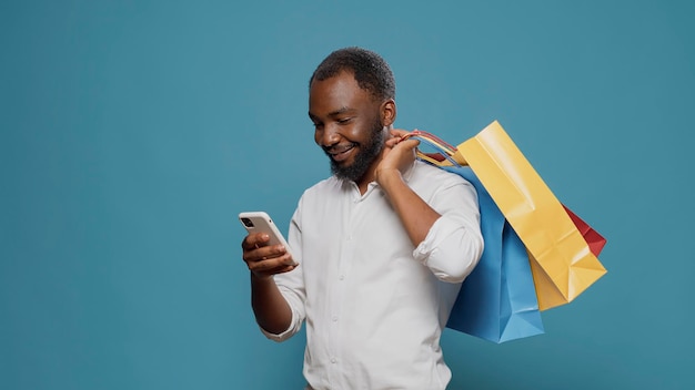 Modern person carrying shopping bags and using smartphone, after making purchase on sale at retail store. Millennial adult holding paper bags and feeling excited about discount shop.