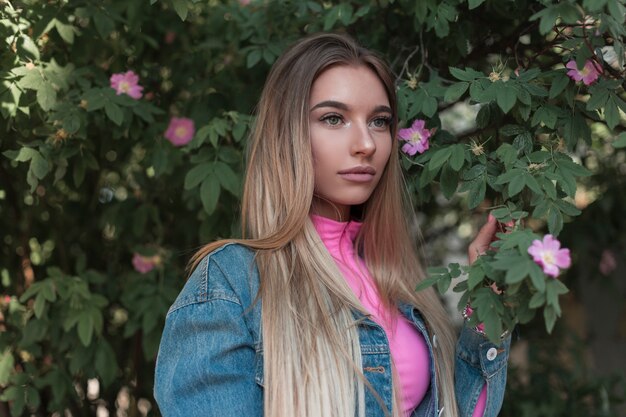 Modern portrait of glamorous young woman in a fashionable denim jacket in a stylish top with beautiful long blond hair relaxes standing near a flowering green bush in the park. Pretty girl model.