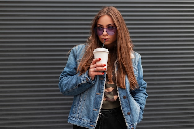 Modern pretty fashionable young hipster woman in stylish violet glasses wearing trendy denim jacket is drinking sweet tasty drink near the metal gray wall in the city. Beautiful girl model outdoors.