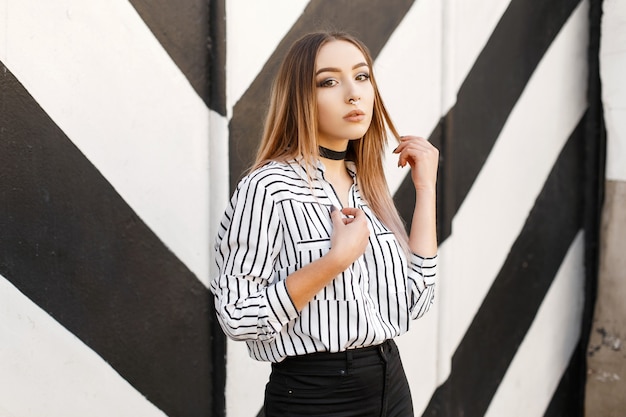 Modern pretty young woman in a trendy black and white blouse in a line with a pierced nose in black jeans in a vintage velvet necklace posing near a striped vintage wall.