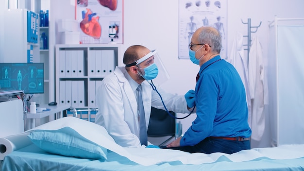 In modern private clinic doctor is listening heart of senior patient sitting on hospital bed. Healthcare medical medicine consultation, using stethoscope giving professional treatment to sick ill diag