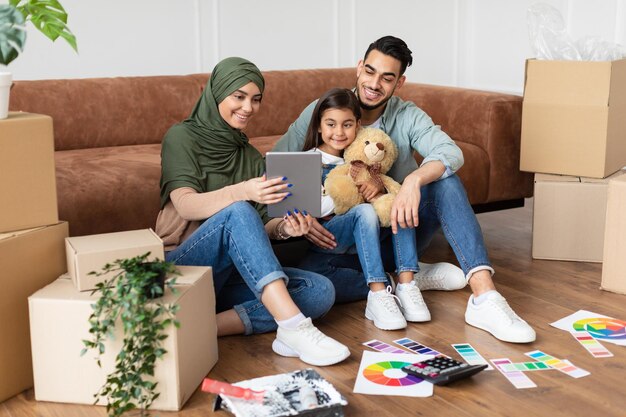 Modern Remote Communication Concept. Portrait of positive arabic family making virtual video call with friends or grandparents using tablet, sitting on floor carpet with paint, talking online