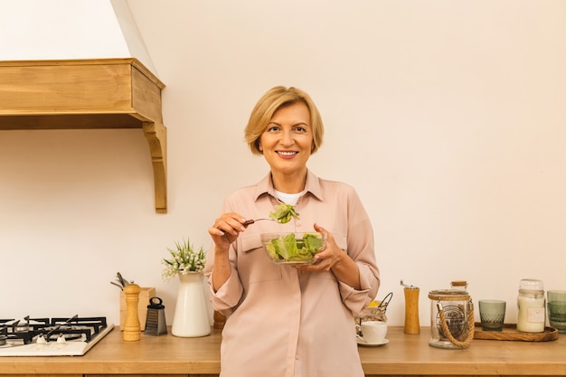 Modern senior aged mature woman eating fresh green salad and vegetables in kitchen, smiling happy. Helthy lifestyle concept.