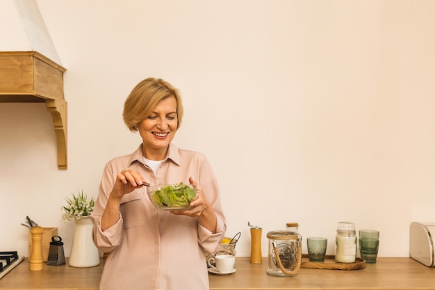 Modern senior aged mature woman eating fresh green salad and vegetables in kitchen, smiling happy. Helthy lifestyle concept.