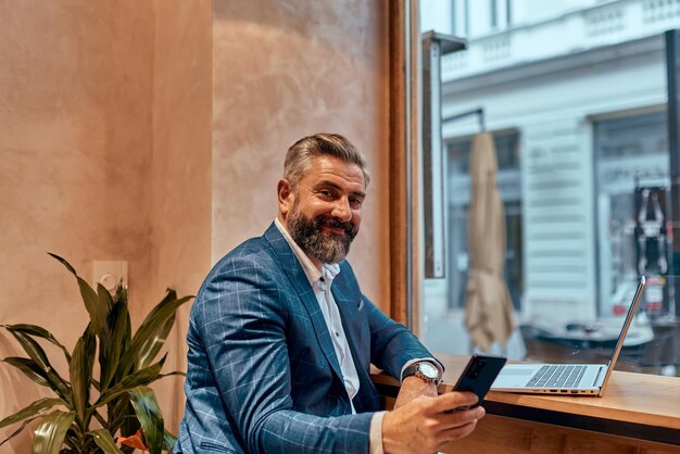 Modern senior businessman sitting in cafe and using laptop and smartphone.