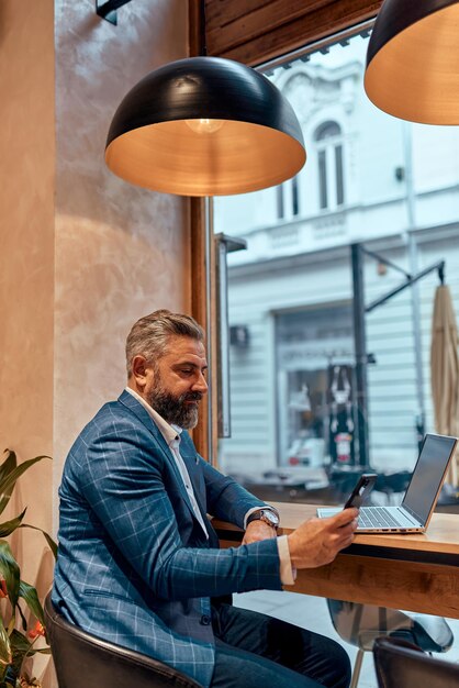 사진 현대의 원로 사업가는 카페에 앉아 노트북과 스마트폰을 사용합니다.