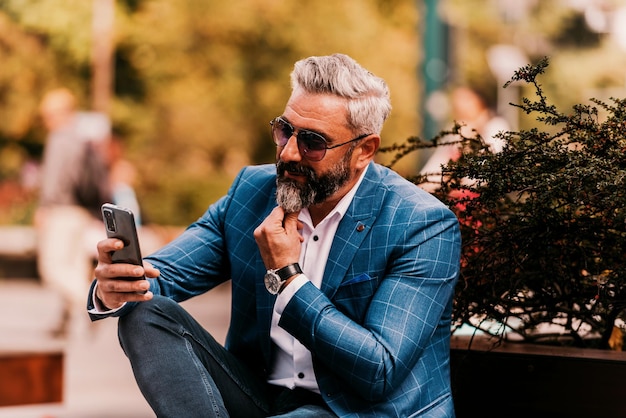 Modern senior businessman using smartphone for online meeting while sitting on bench