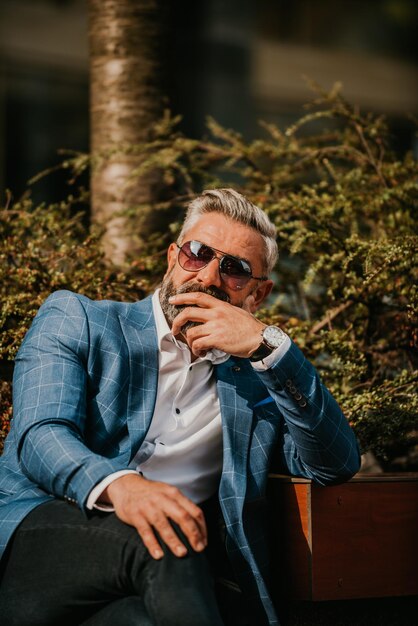 Modern senior businessman using smartphone for online meeting while sitting on bench.