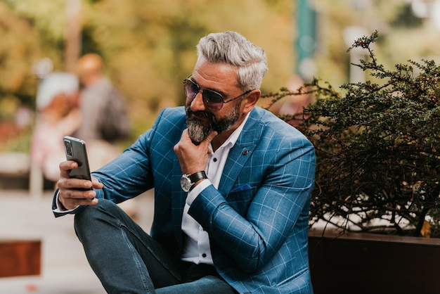 Современный старший бизнесмен использует смартфон для онлайн-встреч, сидя на скамейке.