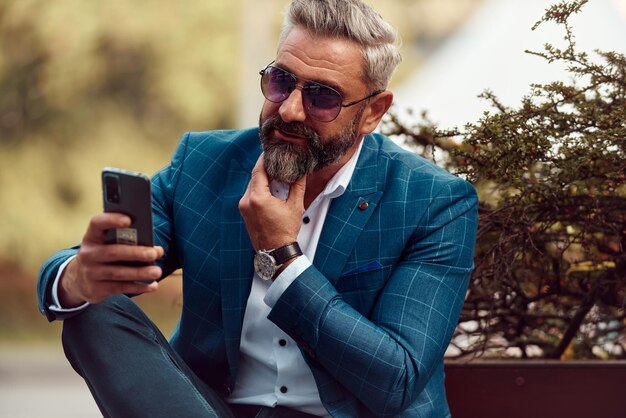 Modern senior businessman using smartphone for online meeting while sitting on bench .