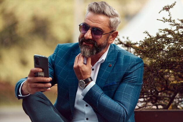 Modern senior businessman using smartphone for online meeting while sitting on bench .