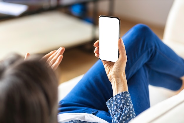 Modern senior woman holding phone with blank screen in hand lying on the couch at home