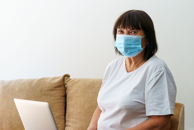 Donna anziana moderna che indossa una maschera medica seduta sul divano di casa utilizzando un gadget per computer felice matura ...