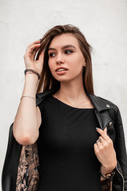 Modern sensual young woman straightens hair. Trendy attractive girl with beautiful eyes with sexy lips in a fashionable leather jacket in a stylish dress posing near a vintage white wall in the city.