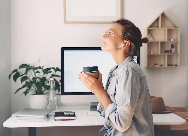 Modern smiling beautiful woman working and drinking coffee from reusable mug at home office Minimalist slow life style workplace of creative female in glasses and earphones using computer technology