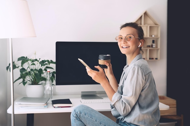 Modern smiling beautiful woman working and drinking coffee from reusable mug at home office Minimalist slow life style workplace of creative female in glasses and earphones using computer technology