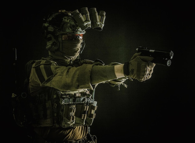 Modern soldier counter terrorist squad fighter in combat uniform helmet and tactical radio headset aiming service pistol in darkness shooting with handgun studio portrait on dark background