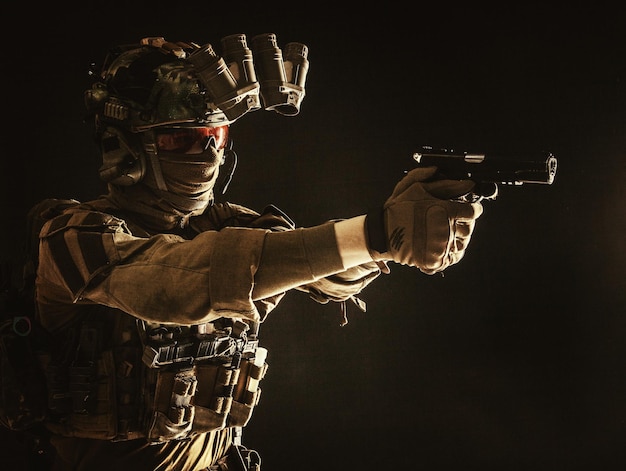 Фото Современный боец контртеррористического отряда в боевой форме, шлеме и тактической радиогарнитуре, нацеленный на служебный пистолет в темноте, стреляющий из пистолета в студийном портрете на темном фоне