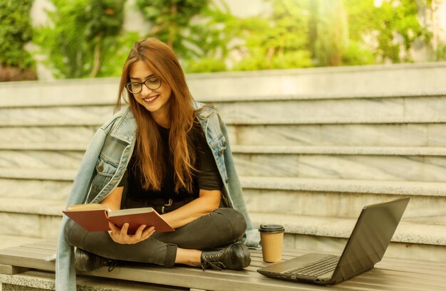 현대 학생. 원격 교육. 젊은 열정적인 여성은 노트북을 들고 벤치에 앉아 책을 읽습니다.