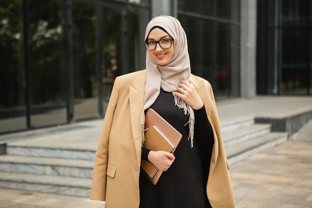 Современная стильная мусульманка в хиджабе, куртке делового стиля и черной абайе гуляет по городской улице с ноутбуком