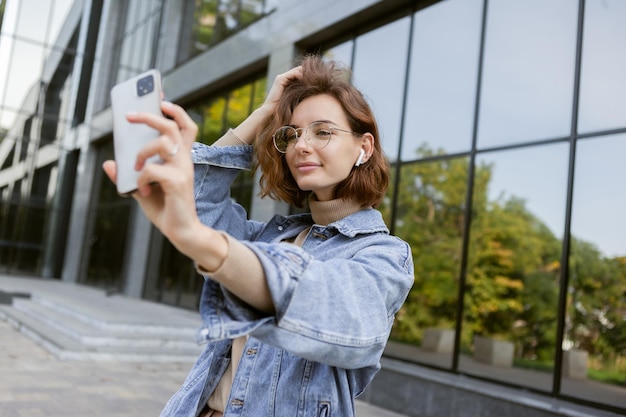 カジュアルな服装でモダンでスタイリッシュな女性は、街の屋外でスマートフォンで自分撮りをします