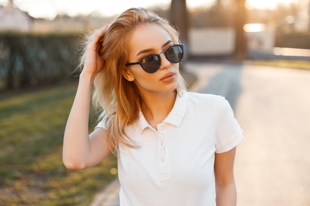 Modern stylish young hipster woman in trendy white polo t-shirt in black sunglasses stand and posing on the street. American beautiful girl.