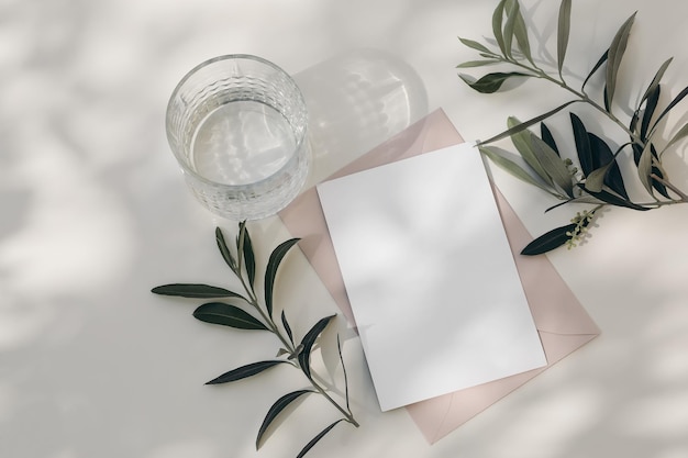 Modern summer wedding stationery still life Drinking glass with shadows Blank blush pink envelope card mock up scene White table background in sunlight Green olive tree branches Flat lay top