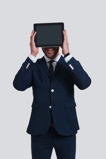 Modern technologies. Good looking young man in full suit covering face with digital tablet and smiling while standing against grey background