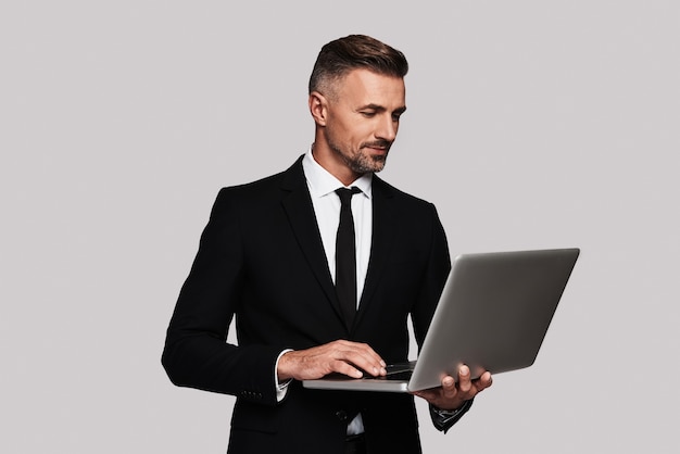 Modern technologies. Handsome young man in full suit using laptop and smiling while standing against grey background