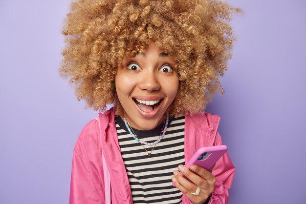 Modern technologies and mobile apps concept positive surprised
woman with curly hair holds mobile phone feels happy chats online
wears striped jumper and pink jacket isolated over purple
background