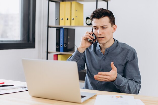 Modern technology and communication concept. Good-looking unshaven young businessman employee working on generic laptop computer and having phone conversation in office