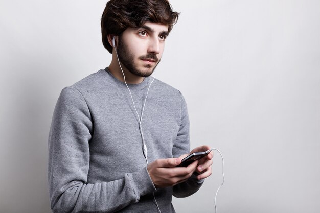 現代の技術とコミュニケーションの概念。彼のスマートフォンを保持している白いイヤホンでひげを持つハンサムな流行に敏感な男の子