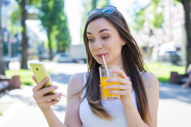 Modern technology teen concept. Closeup photo portrait of funny funky humorous fooling grimacing comic lady holding using telephone cellular gadget device in hands enjoying orange beverage