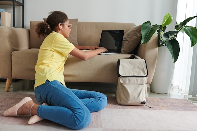 Modern tienermeisje met oordopjes en bril zittend op de bank en typend op laptop terwijl ze huiswerk maakt met behulp van online service