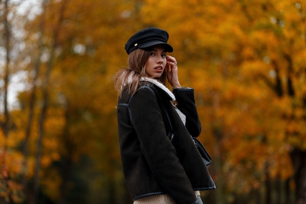 森の中の金の葉を背景に暖かい茶色のスタイリッシュなジャケットのバイザーと黒い帽子の現代のトレンディなヨーロッパの若い女性。ファッショナブルな女の子。女性のカジュアルアウターの新しいコレクション