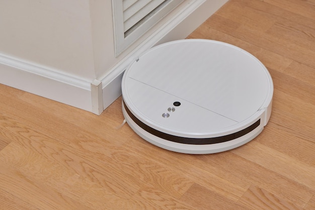 A modern white robot vacuum cleaner in the corner of the\
kitchen room cleans the tiled floor