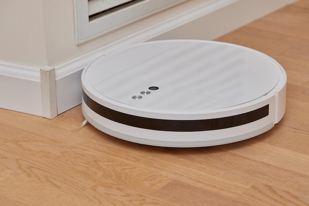 A modern white robot vacuum cleaner in the corner of the\
kitchen room cleans the tiled floor