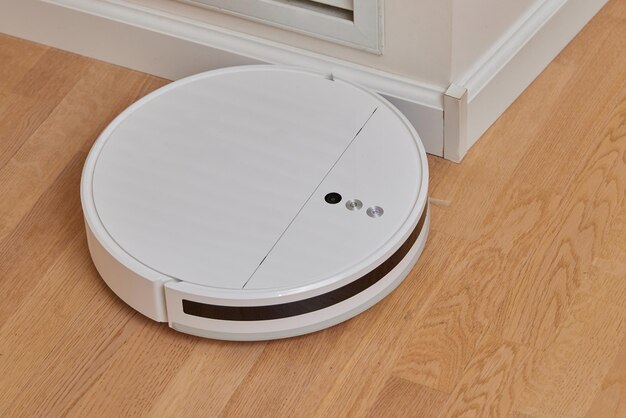 A modern white robot vacuum cleaner in the corner of the kitchen room cleans the tiled floor